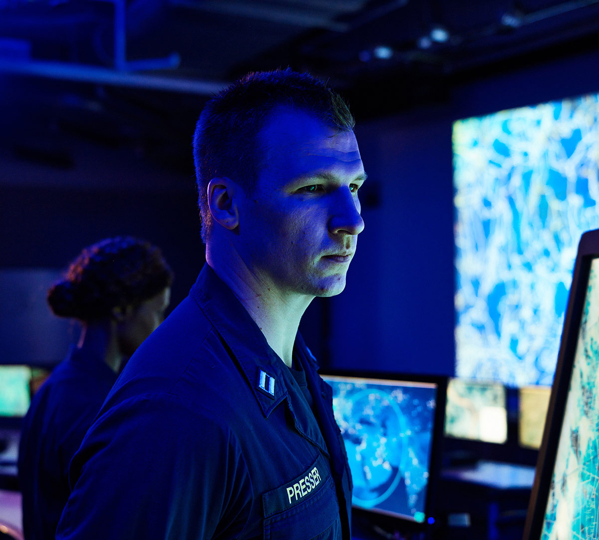 white man in blue military uniform in front of computer monitors
