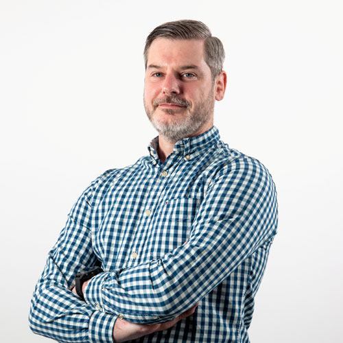 Photo of man with short hair and beard in checked shirt with arms crossed smiling