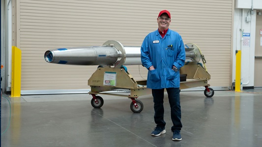 Man wearing blue jacket and red hat standing in front of a rocket.