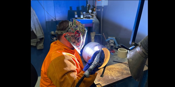 Thomas wears an orange safety uniform and a colorfully graffitied face shield while welding at an industrial workstation