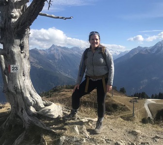 Woman dressed in outdoor gear on a hiking trail surrounded by mountain scenery.
