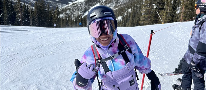 Person in purple overalls, helmet and goggles standing on a mountain top covered in snow.
