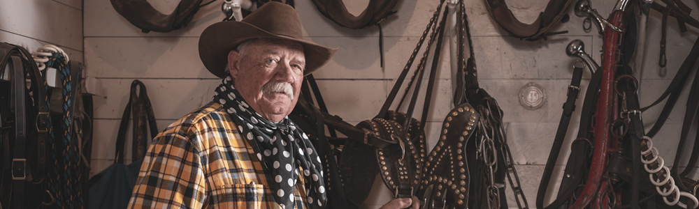 Boyd_Northrop_Grumman_Ranch_Promontory_Utah Older Caucasian man in cowboy attire poses in front of horse saddles hanging on wall