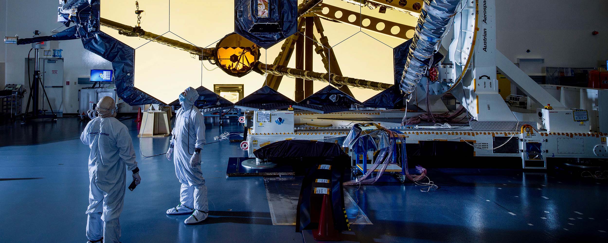 Creating Zero G on a Planet Weighed Down by Gravity Banner Two people in clean suits look up at the James Webb Space Telescope