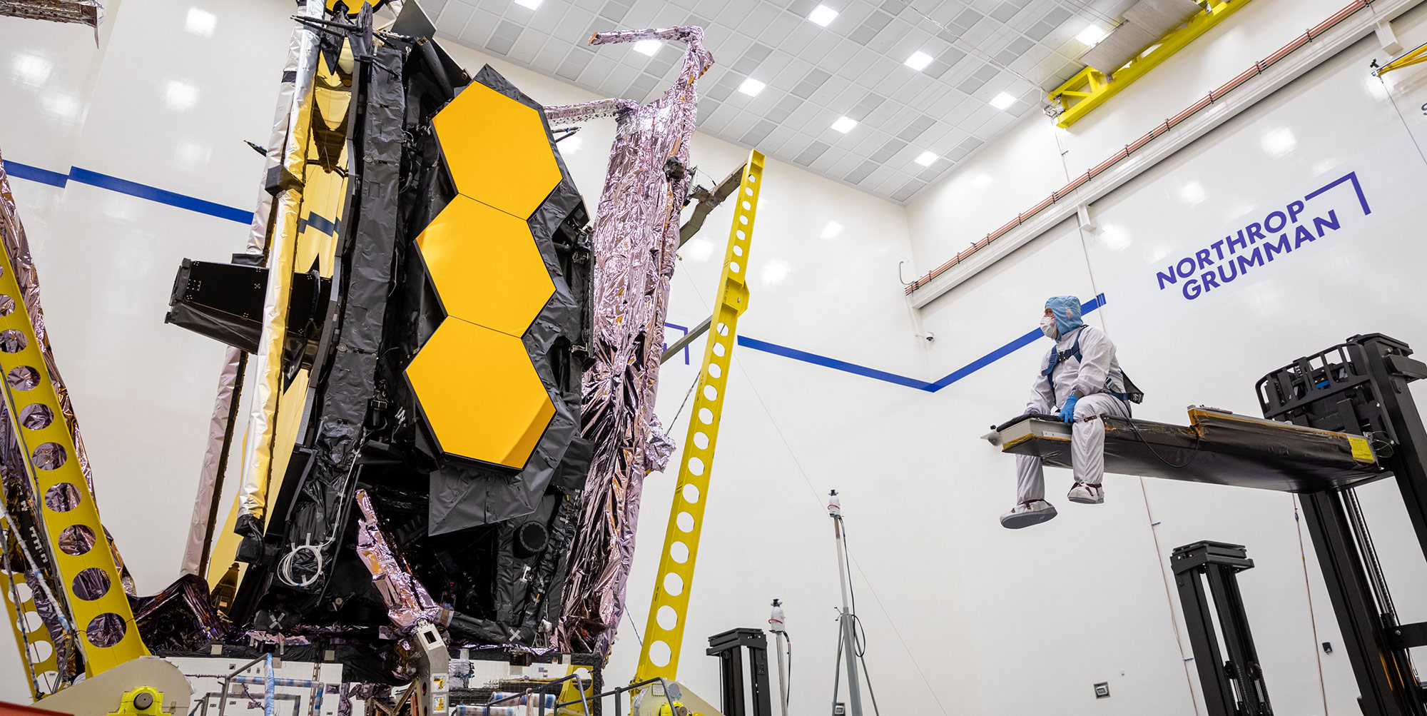 Digital Modeling for Out-of-This-World Environments An engineer in a clean suit sits on a crane and looks at the James Webb Space Telescope sunshield