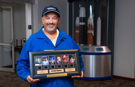 A Latino man in a blue work suit and cap holding a plaque