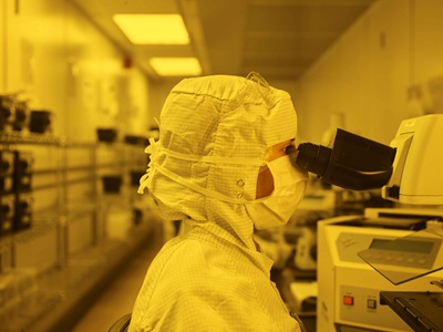 Woman wearing PPE in a clean room with a yellow tint, looking into microscope.