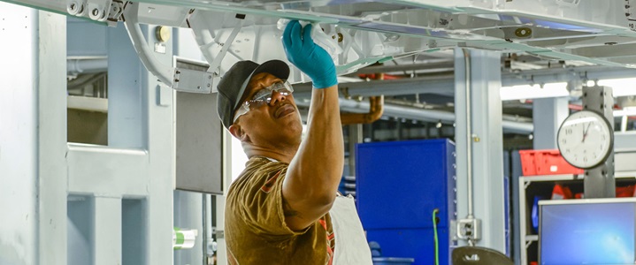 Using Automation and Robotics in Advanced Aircraft Production A man wearing gloves, a hat, safety glasses and an apron works on a machine on Northrop Grumman's Integrated Assembly Line